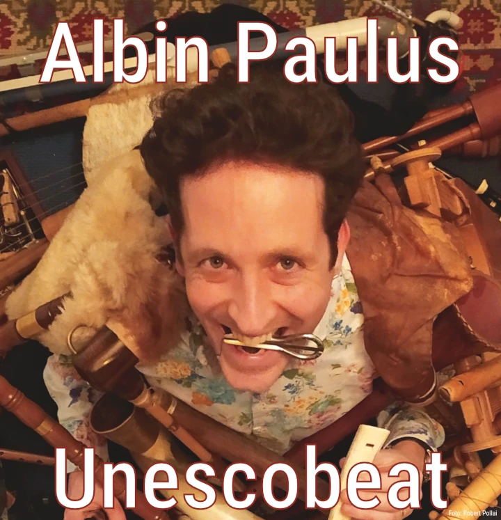 Albin Paulus, von oben fotografiert. Er hält eine Maultrommel zwischen den Zähnen und ist umgeben von vielen auf dem Boden liegenden Musikinstrumenten